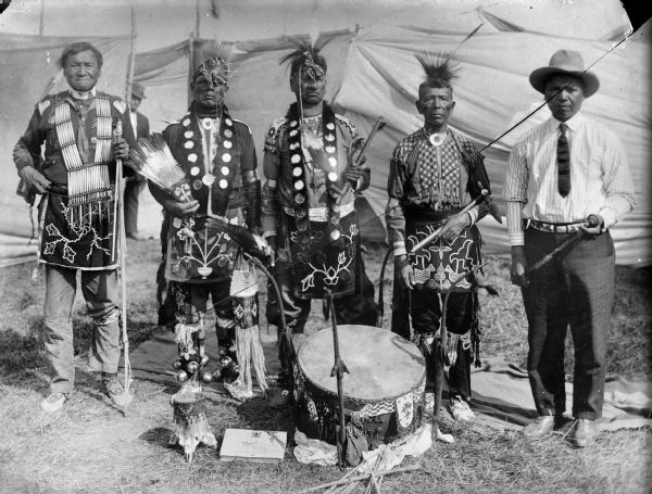 Copy photograph of five Ho-Chunk men posed standing behind a Dream Drum in front of cloth barriers or tents. A powwow group from the 1908 Homecoming, identified from left to right as: Jim Swallow, William Massey (Massie), Thomas Thunder, George Eagle, and Ben Thundercloud. The last three were reportedly listed as full brothers on the 1881 tribal rolls. They are all dressed in regalia, except for Ben Thundercloud, who is wearing a shirt, tie, and hat.