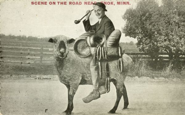 An older man is riding a sheep on a dirt road. He has a small barrel, some sausage links, and a loaf of bread. The object in his right hand appears to be a crop. A fence protects a pasture in the background.  The image was pieced together from a number of sources; a close look betrays that the man's legs are fake. Red text in the upper portion bears the inscription "Scene on the Road near Enok, Minn."