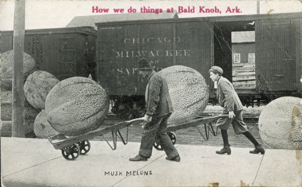 A man and a boy use dollies to haul giant musk melons near a train. In red text on the upper center portion is the inscription, "How we do things at Bald Knob, Ark," suggesting that this postcard, though photographed in Waupun, was distributed as an image of Bald Knob, Arkansas. The train in the background reads, "Chicago, Milwaukee and Saint Paul".