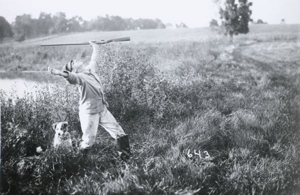 Photographic mock-up for an Alfred Stanley Johnson photomontage postcard entitled "Kickers." A boy wearing waders and holding a rifle stands with his arms outstretched and his face toward the sky. A dog is by his side; the two are standing by the shore of a pond.
