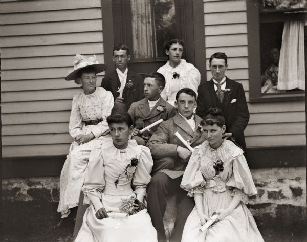 Eight men and women of a graduating class sit outdoors holding their diplomas. They are seated on the steps of the photographer's home at 131 East Montello Street.