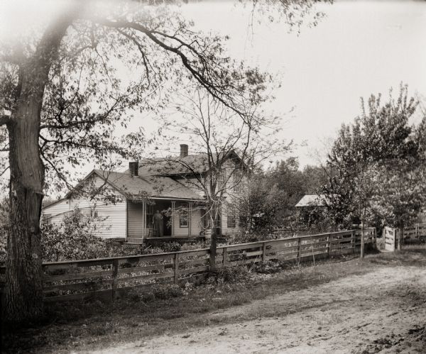 View from road towards a man standing on the porch of a home belonging to either Mrs. Kelly or Mrs. Stevenson. A fence runs along the road in front of the yard and house.