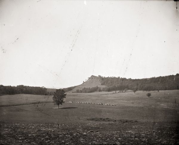Distant view of Black Hawk Lookout near the Wisconsin River. There is an open field with a single tree and a fence in the foreground.