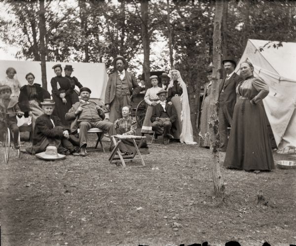 Group of campers near pitched tents in the woods. Boy on tricycle left side of frame, and women in carriage behind group of four at center.