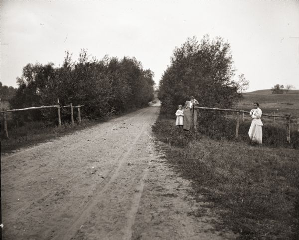 Ada Bass, the photographer's wife, leaning against a fence, next to daughter Everetta Bass as a young girl, along a dirt road. The photographer's sister, Rhoda Bass (later Mrs. William Shephard), is at far right.