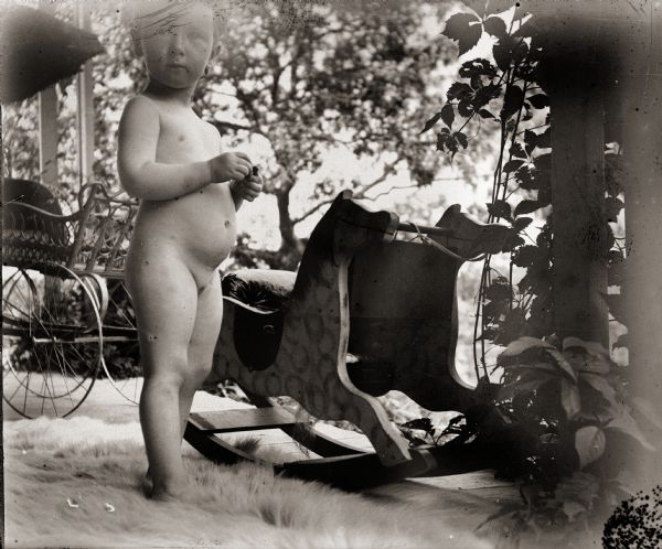 Everetta Bass, the photographer's daughter, standing nude next to rocking horse. She, along with the rocking horse and a baby carriage, are on an unidentified front porch.