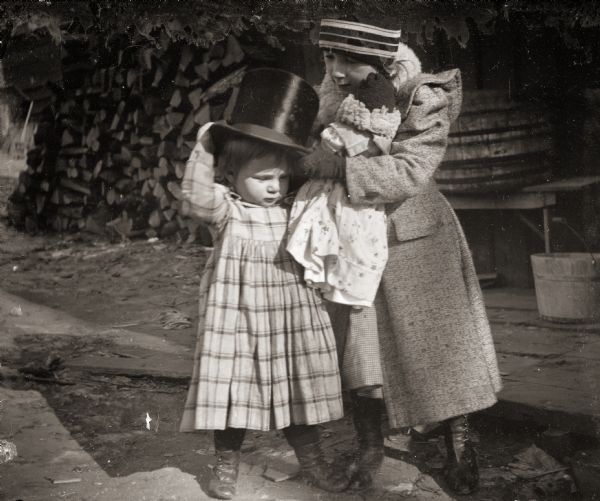 Everetta Bass, the photographer's daughter, helping her brother Cary put on a silk top hat. She is holding a doll and they are standing outside near a firewood pile.