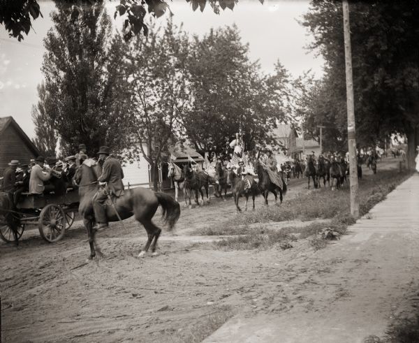 Parade with costumed men on horses, a band being pulled in a wagon and American flags.