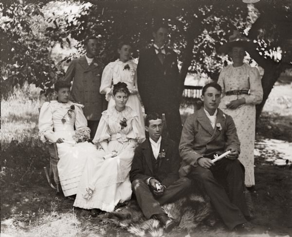 Group portrait of the graduating class. The four men and four women are seated in the front yard of the photographer holding their diplomas.