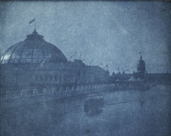 A cyanotype view of the Horticulture Building at the World's Columbian Exposition with the dome of the Illinois Building in the background.  There is a motor launch on the lagoon.