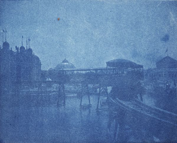 A cyanotype view of the World's Columbian Exposition grounds from near the North Loop elevated railway station, showing the glass dome of the Horticulture Building in the distance and the railroad trestle in the foreground. The Agriculture Building is on the left, and one of the domes of the Fisheries Building is on the far right.