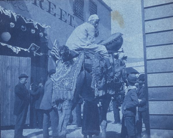 A cyanotype view of Cairo Street on the midway of the World's Columbian Exposition. An Egyptian with a large drum rides a camel as men and boys watch. The man and camel were part of an "Arab wedding procession" performed daily for spectators.