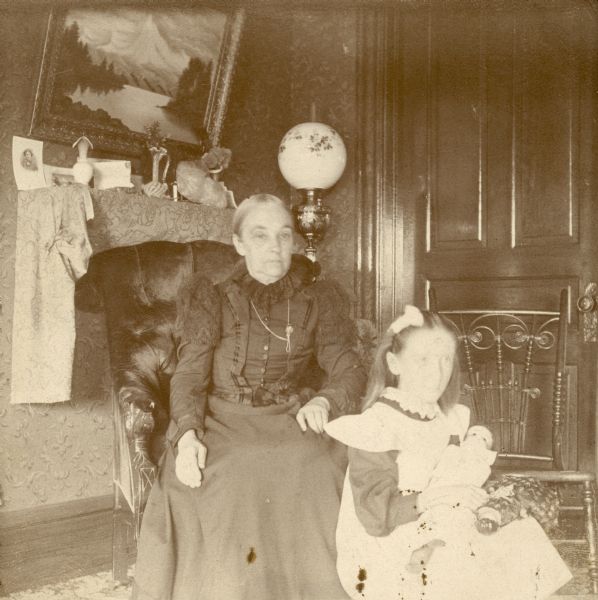 Lorinda Hill Bass and her granddaughter, Everetta Bass, sitting in a parlor. Everetta holds two dolls in her lap. There is a large oil lamp in the background.