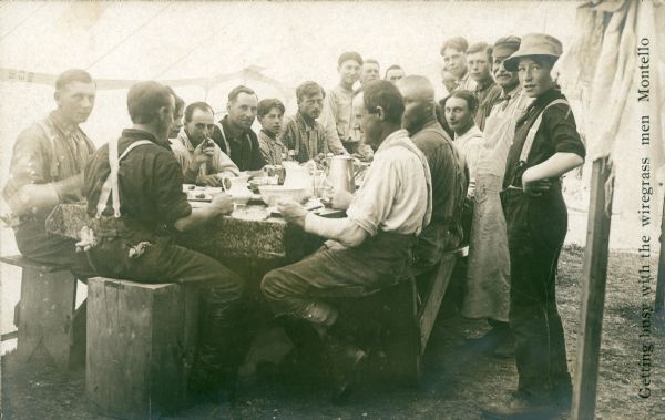 Photographic postcard of a group of men and boys gathered for a meal in a tent. There are benches along the sides of the table. One man sits on a wooden box at the end. There are pitchers, a bowl and metal coffee pot on the table. The cook, second from right, is wearing a cap and long checkered apron. The caption reads, "Getting busy with the wiregrass men Montello."