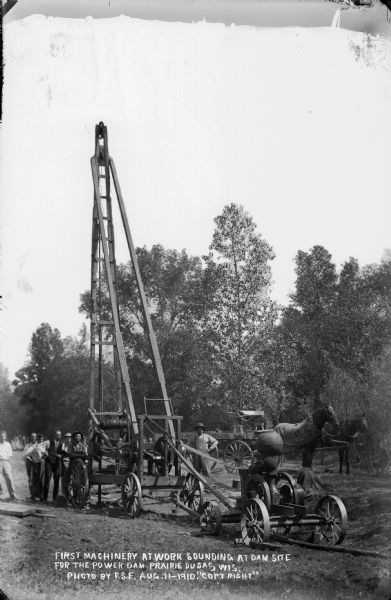The first machinery at work sounding at the site for the power dam.  Workmen are posing near the carriage-mounted equipment. A horse-drawn wagon is in the background.