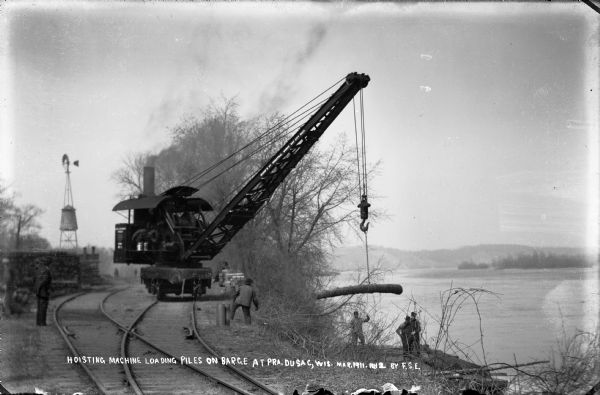 Workers use a rail car mounted crane to load pilings onto a barge.  Bluffs are in the distance.