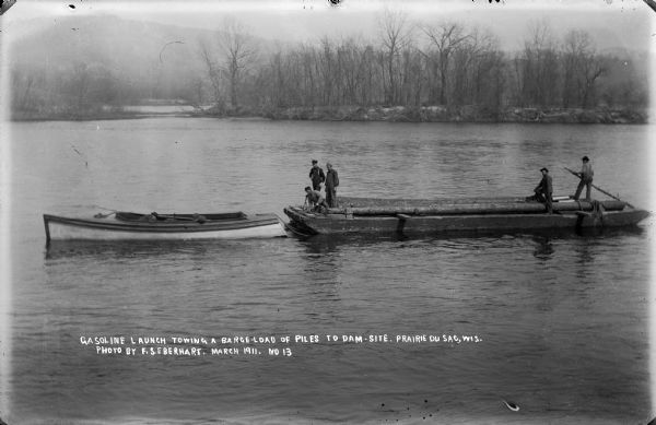 A gasoline-powered launch tows a barge loaded with log pilings on the Wisconsin River to the Prairie du Sac dam site. Workmen drive the launch and guide the barge.