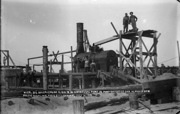 Workmen pose around the "big boiler" and engine that power the 12 inch centrifugal pump used to pump out the coffer dam at the power dam construction site.
