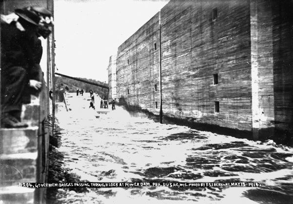 A view upstream through the lock at the Prairie du Sac dam. Workmen are guiding a barge through the lock as a man and woman are watching from steps in the wall in the foreground on the left.