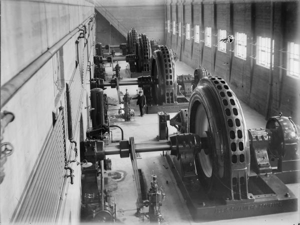 Elevated view of a man standing near the generators inside the power house at the Prairie du Sac dam. The base of the generators is marked with the name of the manufacturer, the Allis-Chalmers Company.