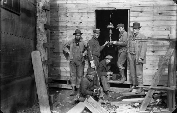 Workers posing in a doorway at the Prairie du Sac dam construction site. Two men appear to be working on a gas lamp, while others are holding spools of wire, preparing for the production of electric power at the site. There is a well-dressed gentleman, possibly a foreman, on the right.