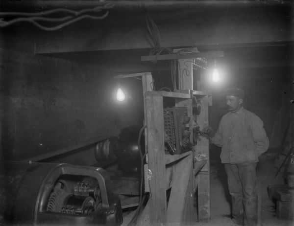 A worker standing at the controls of a generator, most likely in the temporary power house at the Prairie du Sac dam site. The steam driven generator produced power for the electric railroad and other uses during the construction of the dam.