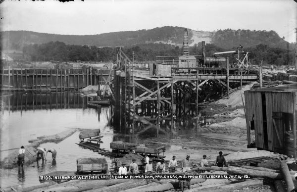 Workers posing inside the coffer dam. The narrow gauge railroad is behind them. One of the pile drivers is in the background. A bare area on the bluff in the background marks the site of the gravel quarry.