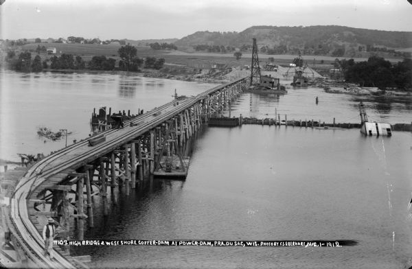 Elevated view of the power dam construction site from the west, showing the elevated trestle, coffer dam, and steam pile drivers. The concrete plant is visible on the east bank. Note that high water has flooded the interior of the coffer dam.