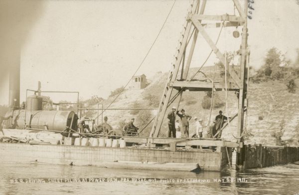 Men posing on a barge carrying a steam powered pile driver at work on the coffer dam for the construction of the powerhouse. Caption reads: "Driving Sheet Piling at Power Dam, Pra. du Sac, Wis."
