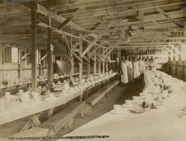 Eight cooks posing inside the dining hall. Long tables are set for a meal.