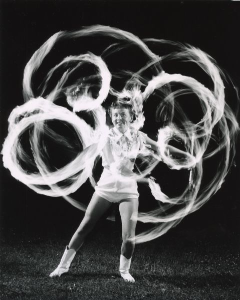 Outdoor photograph of a young woman twirling batons with flaming ends.