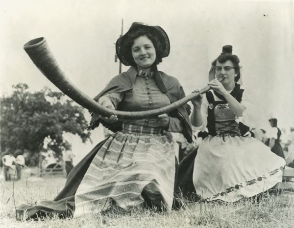 Two women in Swiss costume kneeling in the grass during the Swiss Cheese celebration. One is blowing into an alpine horn while the other holds the instrument.