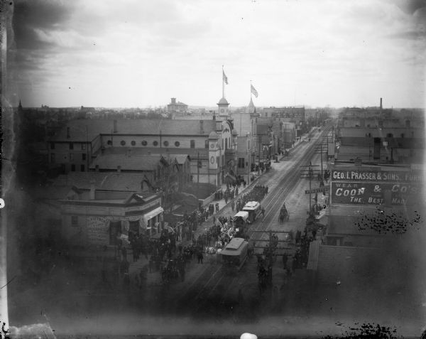 Elevated view looking west on W. National Avenue from about S. 6th Street, towards a parade marching down National Avenue with a crowd of people watching, and signs and flags on surrounding buildings. The ornate structure near the center is probably South Side Turn Hall, which stood between S. 7th and S. 8th Streets.