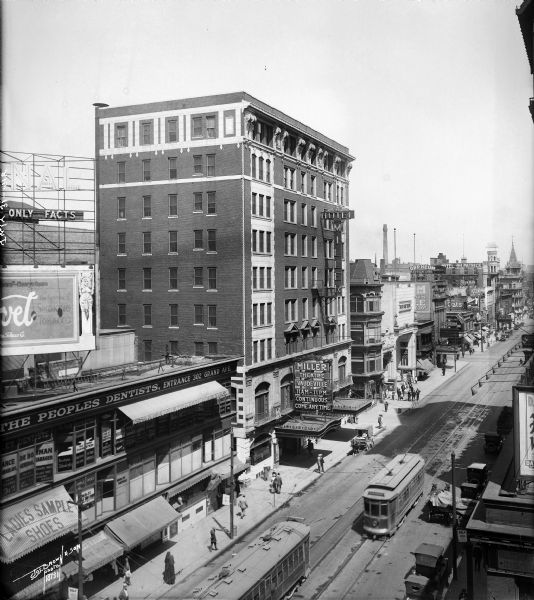 Elevated view looking north on N. 3rd Street from W. Wisconsin Avenue towards the commercial district surrounding the Miller Hotel and Theater. There are streetcars, horse-drawn vehicles, and automobiles on the street, in addition to numerous pedestrians.