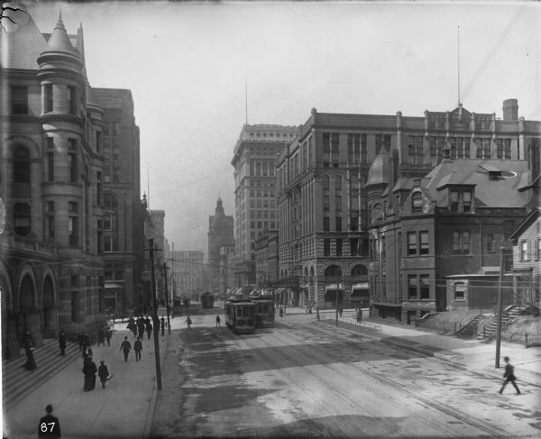 East Wisconsin Avenue looking west down W. Wisconsin Avenue from Jackson Street. The round turret on the left is on the Federal Building, with the Pfister Hotel and Milwaukee Club on the right. On the street are streetcars and automobiles, and numerous pedestrians.