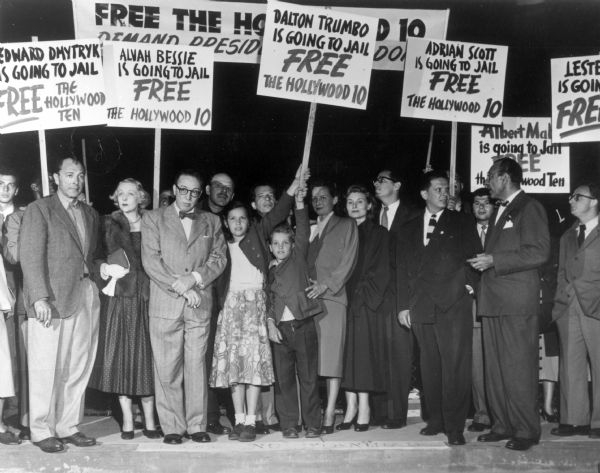 Several members of the screenwriters and directors known as the Hollywood Ten, together with members of their families at a demonstration before their imprisonment for failure to testify before the House Un-American Activities Committee about their Communist Party memberships or associations. On the left with open collar is Lester Cole. Dalton Trumbo stands nearby wearing eyeglasses and a bowtie, with Alvah Bessie looking over his shoulder. Herbert Biberman, also wearing a bow tie, stands second from the right.