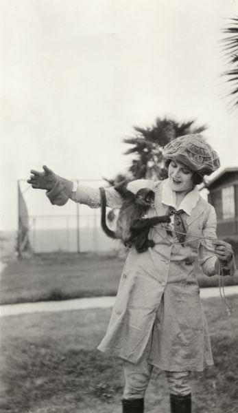 Silent film actress Ruth Roland in riding costume and a velveteen cap playing with a small monkey.