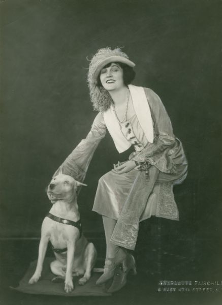 Broadway actress Eleanor Woodruff, wearing a dress with flared sleeves, a feathered hat, and pearls, posed with a white bull terrier.

Original caption:
"Eleanor Woodruff in <i>Back to Methuselah,</i> Garrick Theatre."