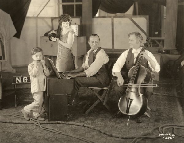 Publicity still for <i>All Souls' Eve</i>, a Realart silent film released in 1921. From left to right: the child actor Mickey Moore plays a harmonica, the star Mary Miles Minter plays a concertina, their director Chester Franklin plays a portable pump organ (harmonium), and an unidentified musician plays the cello.