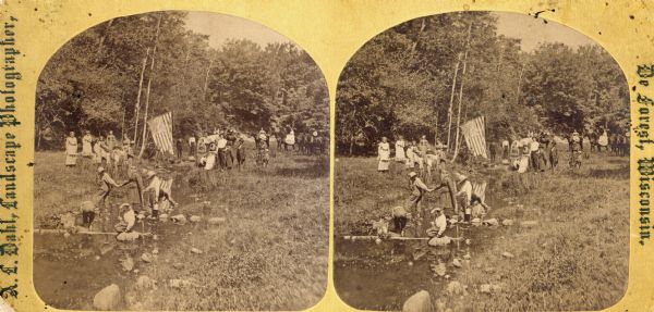 Identified as " Lover's Retreat, Spring View." Group in field with stream on a picnic. Children are stepping on stones in foreground with U.S. flag, people in carriage and people on horses in background.