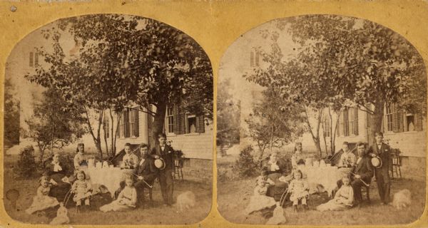 Stereograph of Pastor Claus Friman Magelssen (1830-1904), his second wife Marie Schlambusch Magelssen, and their family sit around a table set for coffee. The parsonage and the Luther Valley Church, built in 1872, are behind them. Visible are the minister's pipe, a baby carriage, a girl's doll and a dog.