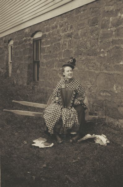 Emma Olson, member of the Menomonie High School class of 1905, depicted as a pauper. Pictured sitting in a wheelbarrow, wearing tattered clothes, and playing the accordion. Part of a yearbook created by classmate Albert Hansen, based on a class prophecy theme.