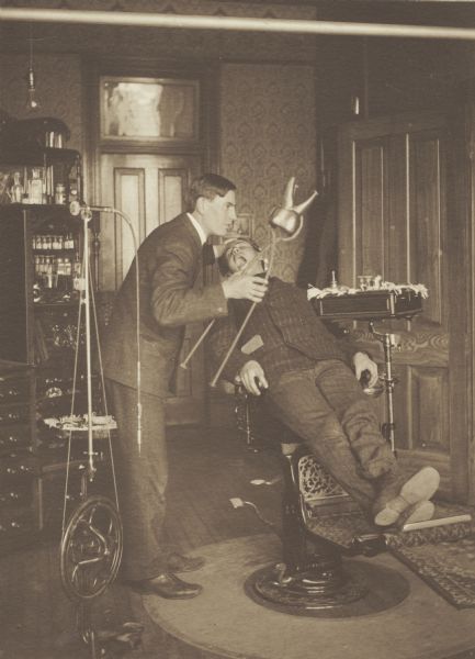 Frank William Kelley, member of the Menomonie High School class of 1905, depicted as a dentist. He is pictured holding a large dental apparatus and a large tooth, and is standing over a patient. Part of a yearbook created by classmate Albert Hansen, based on a class prophecy theme.