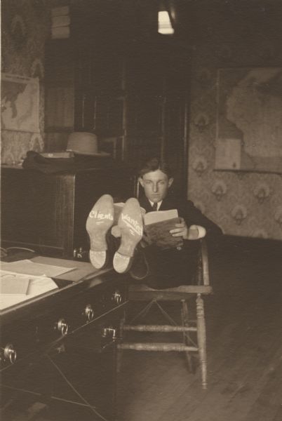 Paul H. Ryan, member of the Menomonie High School class of 1905, depicted as an attorney, sitting at a desk with his feet up. The bottom of his shoes bear the words, "Clients Wanted." Part of a yearbook created by classmate Albert Hansen, based on a class prophecy theme.