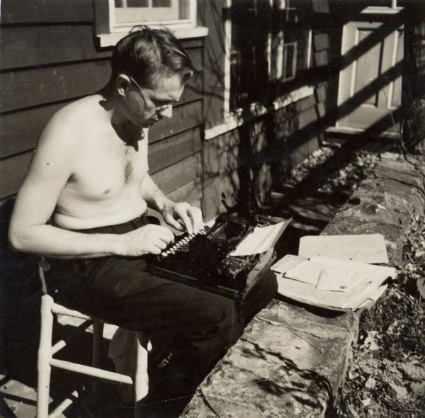 Myles Horton sitting, shirtless, typing on a typewriter on his lap while sitting outdoors.