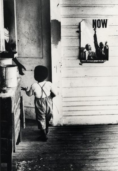 Child Next to SNCC Poster A small African American child walking towards a door where a woman is holding her hand out. There is a SNCC (Student Nonviolent Coordinating Committee) poster on the wall.