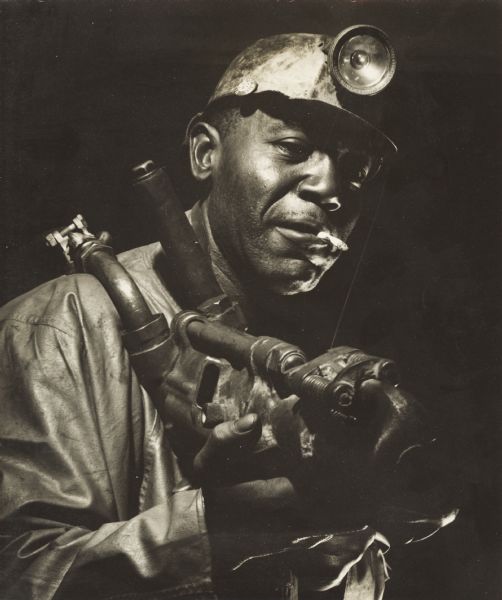 An African American ore miner, holding a large tool and smoking a cigarette. A pin that reads, "Mine-Mill-CIO" is attached to his hardhat.