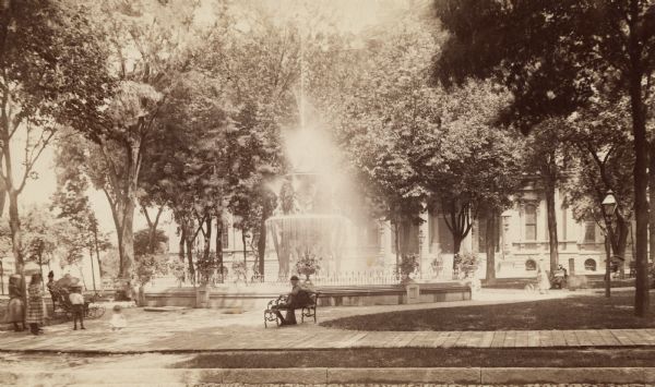 Fountain in center, with trees along the border of the small square. A man sits on a bench and children are on the left. The courthouse is visible in the background.