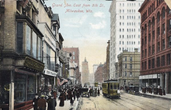 Looking east from 5th Street. Road lined with buildings, and pedestrians crowd the sidewalks. Horse-drawn vehicles and a trolley are on the road. Caption reads: "Grand Ave. East from 5th St., Milwaukee, Wis."