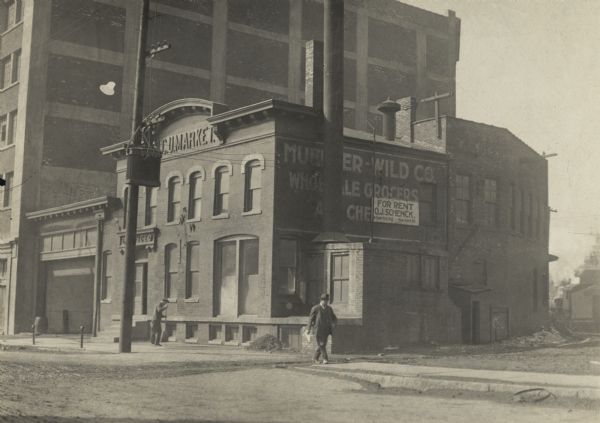 Exterior of building.  A large advertising sign is on the right side, behind a large smokestack.  A smaller sign stating "For Rent O.J. Schenck" has been hung over the larger sign.  On the left is a an open garage attached to the building.